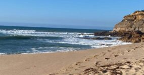Beachside Pilates class on sandy shore with rocky cliffs, ocean waves, and clear blue sky.